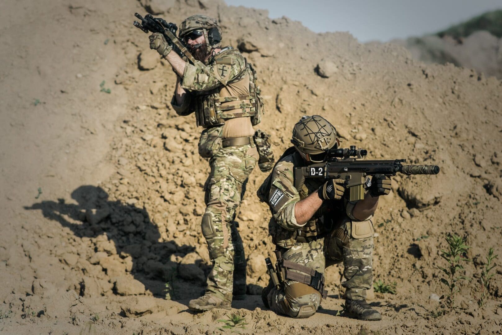 Two soldiers in camouflage are standing and kneeling.