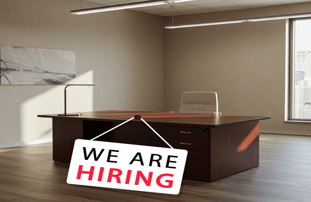Vacant desk and chair in an office space with a sign on front that says We are hiring